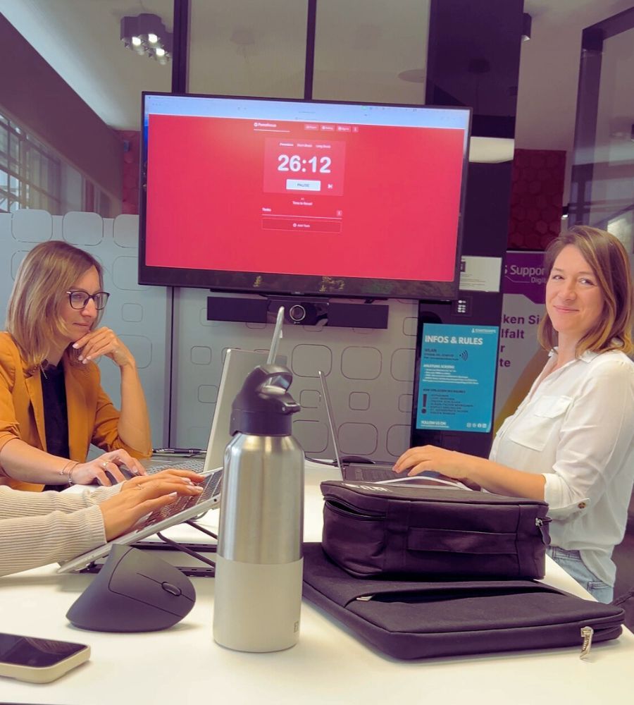 Sarah und zwei weitere Frauen arbeiten mit ihrem Laptop. Im Hintergrund hängt ein Bildschirm der einen Timer von 26:12 Minuten anzeigt